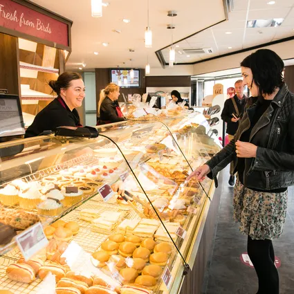 Kunde wählt Gebäck an einer Bäckereitheke mit Kuchen, Brötchen und Brot, während das Personal hinter der Auslage hilft.