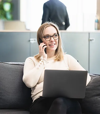 Person sitzt auf einem Sofa, nutzt einen Laptop und spricht am Mobiltelefon in einer hellen modernen Büroumgebung.