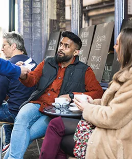 Personen sitzen vor einem Café, während eine Person eine Zahlungskarte an eine andere in der Nähe eines kleinen runden Tisches mit Getränken übergibt.