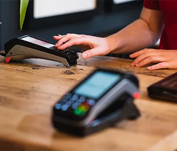 Nahaufnahme einer Person, die ein Zahlungsterminal auf einer Holztheke bedient, mit einem weiteren Kartenlesegerät im Vordergrund.