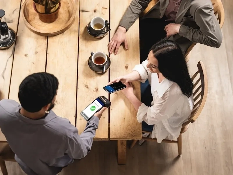 Ein Kellner hält ein kabelloses Zahlungsterminal, während ein Kunde seine Karte am Restauranttisch zum Bezahlen auflegt