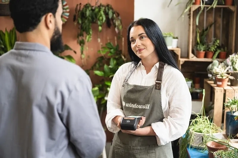 Person mit einer schürze hält ein portables zahlungsterminal und steht einem kunden in einem pflanzengeschäft gegenüber