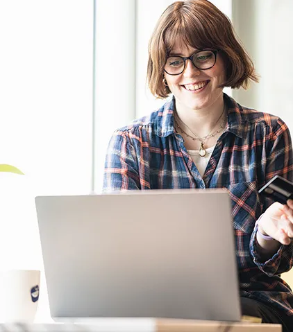 Person mit kariertem hemd nutzt laptop haelt kreditkarte mit tasse auf dem tisch in hellem raum