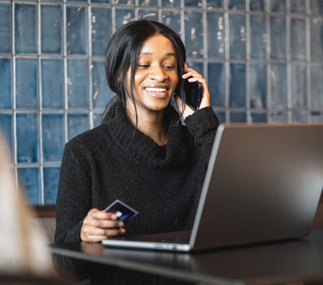 Person hält eine kreditkarte und telefoniert mit einem handy während ein laptop an einem tisch genutzt wird