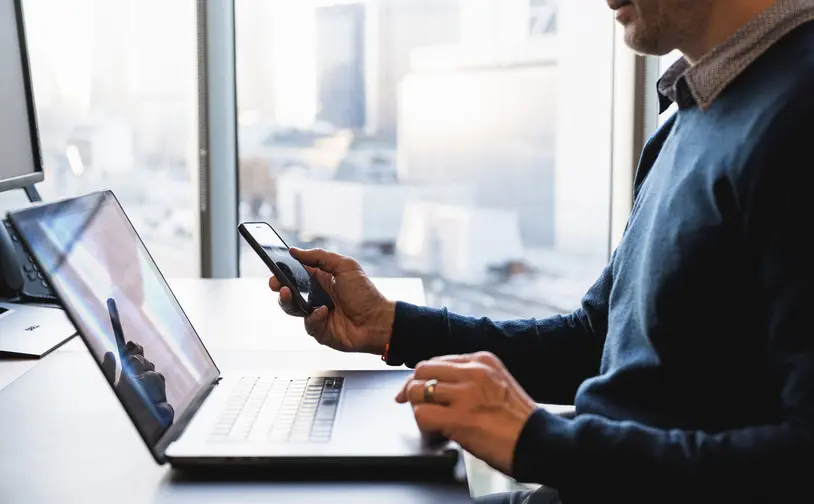Person nutzt einen laptop und hält ein smartphone an einem schreibtisch vor großen fenstern