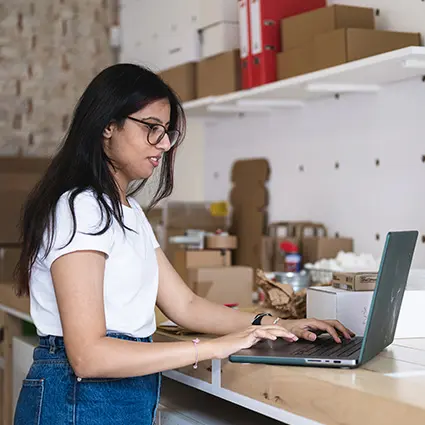 Person steht an einer werkbank und nutzt einen laptop in einem raum mit regalen und kartonboxen
