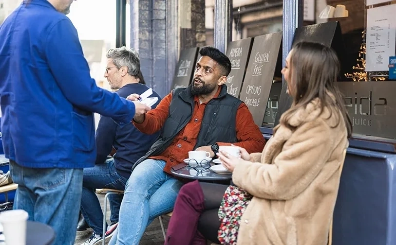 Personen sitzen im außenbereich eines cafés, eine person übergibt einen gegenstand mit speisetafeln im hintergrund