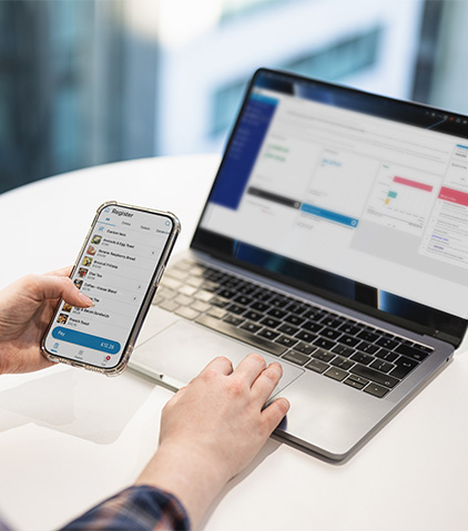 Eine Person verwendet gleichzeitig ein Smartphone und einen Laptop mit einer App, die eine Liste auf dem Telefon und ein Dashboard mit Diagrammen auf dem Laptopbildschirm anzeigt.